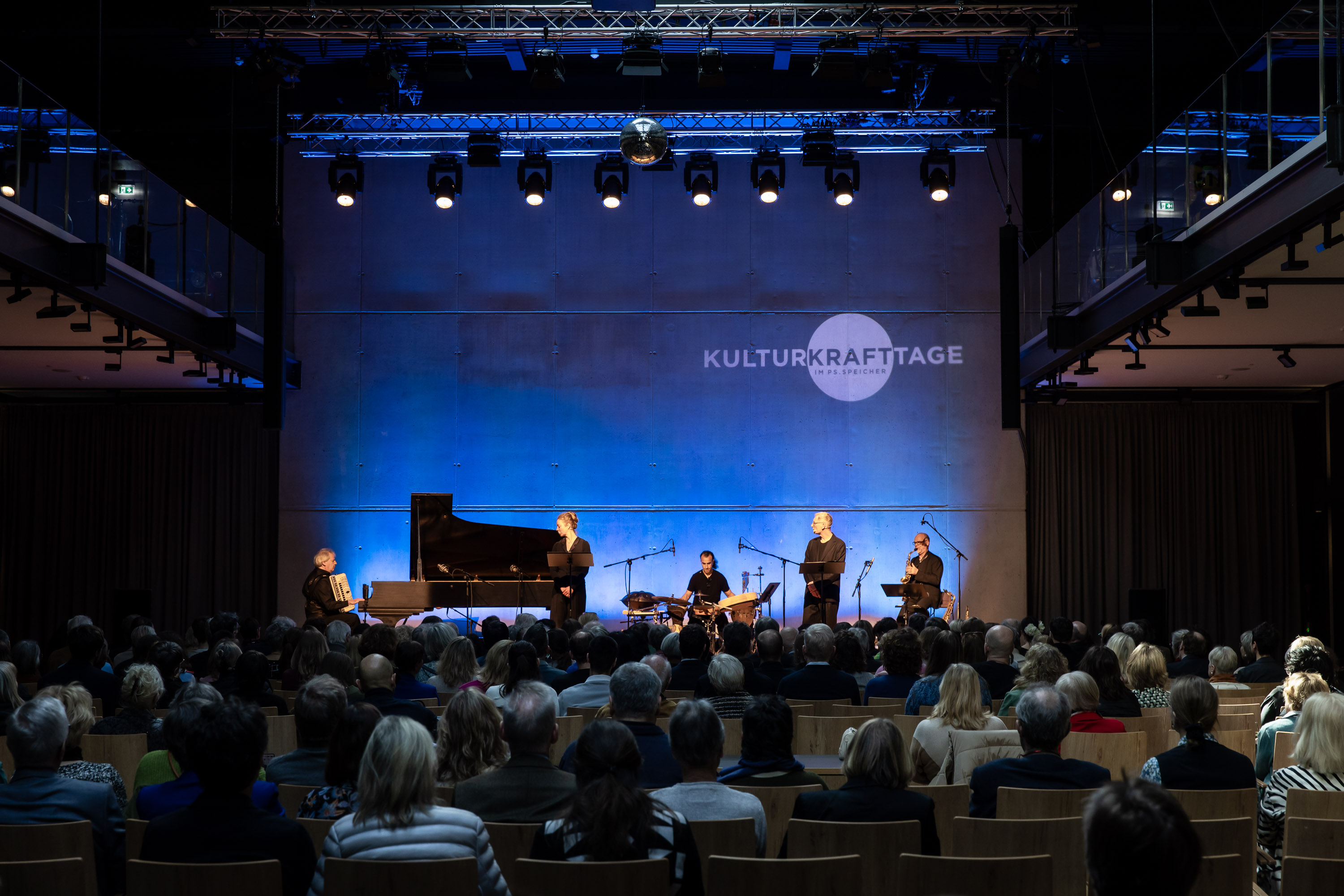 Eine Band bestehend aus Akkordeon, Violoncello und Klarinette spielt vor einem sitzenden Publikum. Alles ist in blaues Licht gehüllt