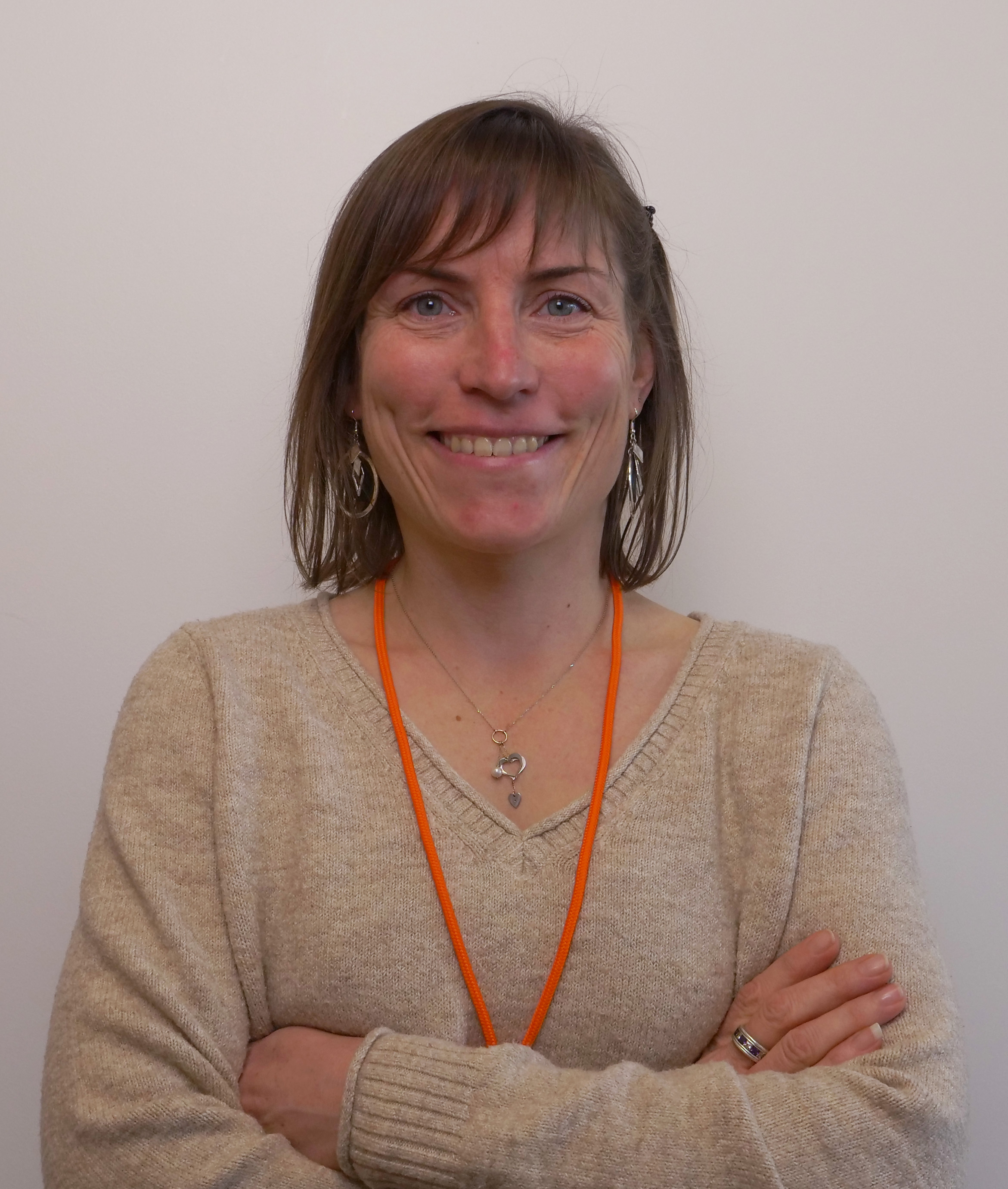 Femme souriante avec les bras croisés, portant un pull beige et un collier. Elle a les cheveux longs et une lanière orange autour du cou.
