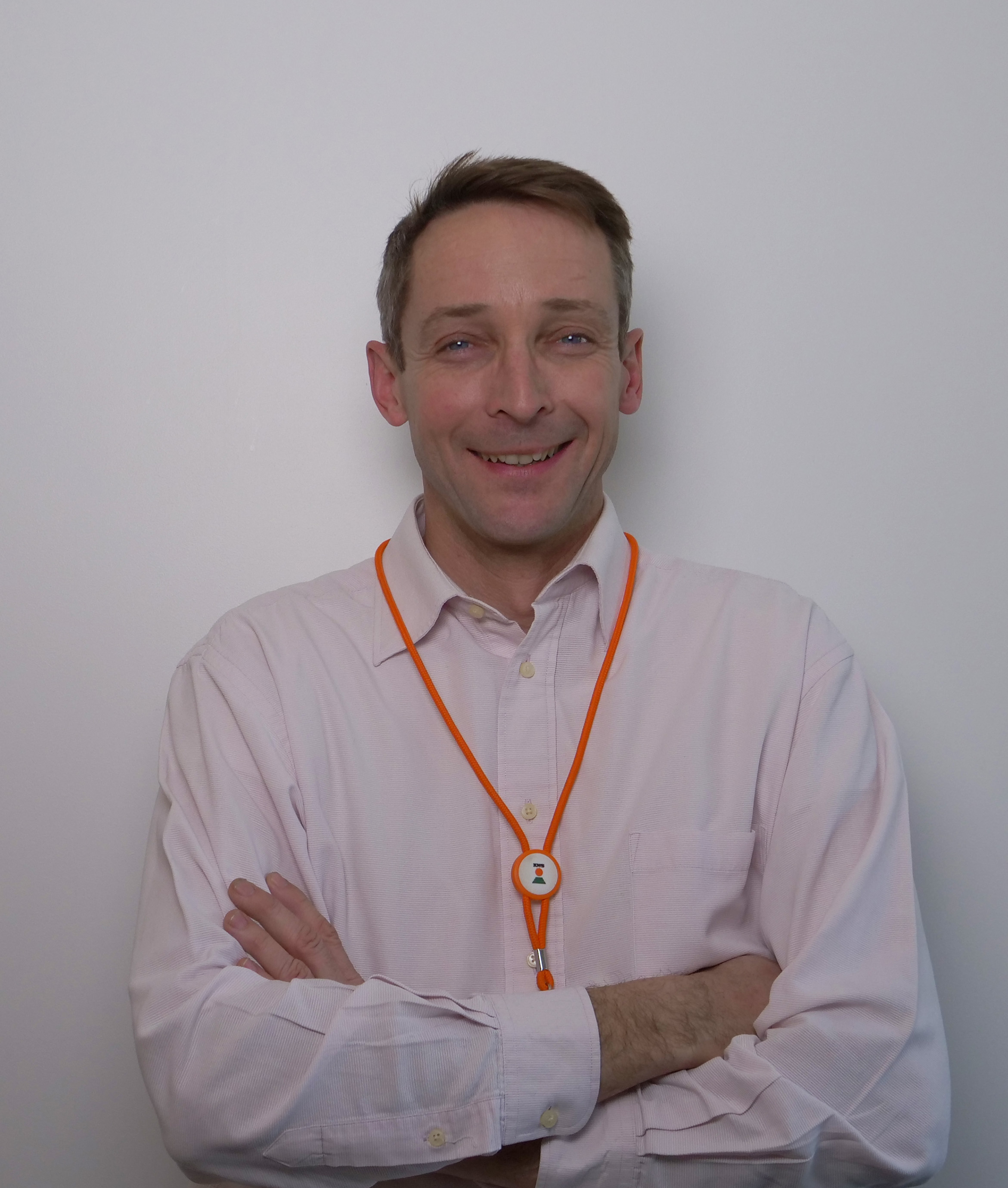 Homme souriant avec un collier orange, les bras croisés, portant une chemise claire. Fond blanc.