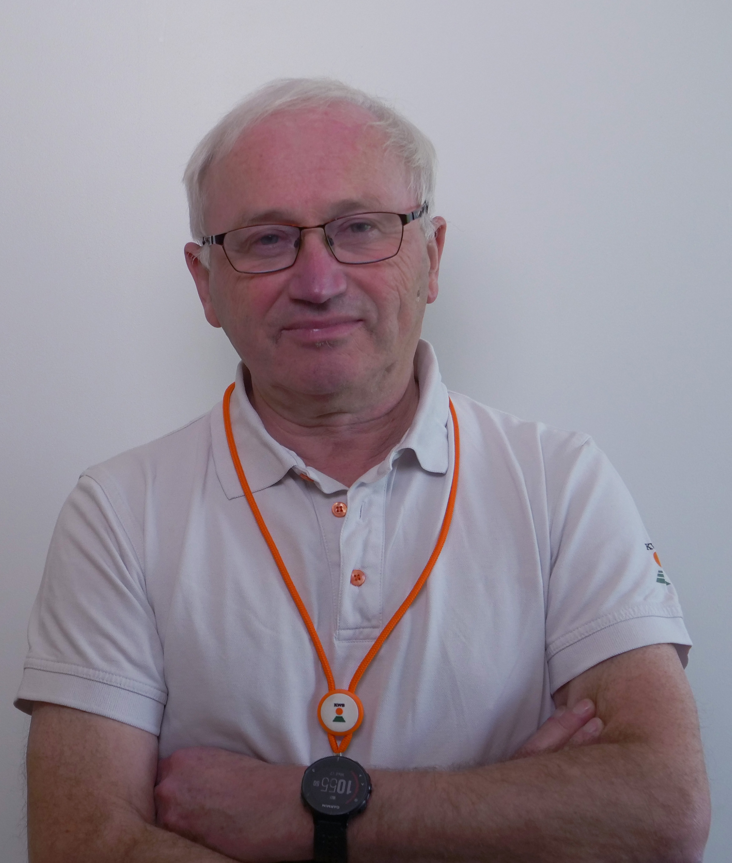 Homme avec des lunettes et un polo blanc, les bras croisés, portant un collier orange et une montre.