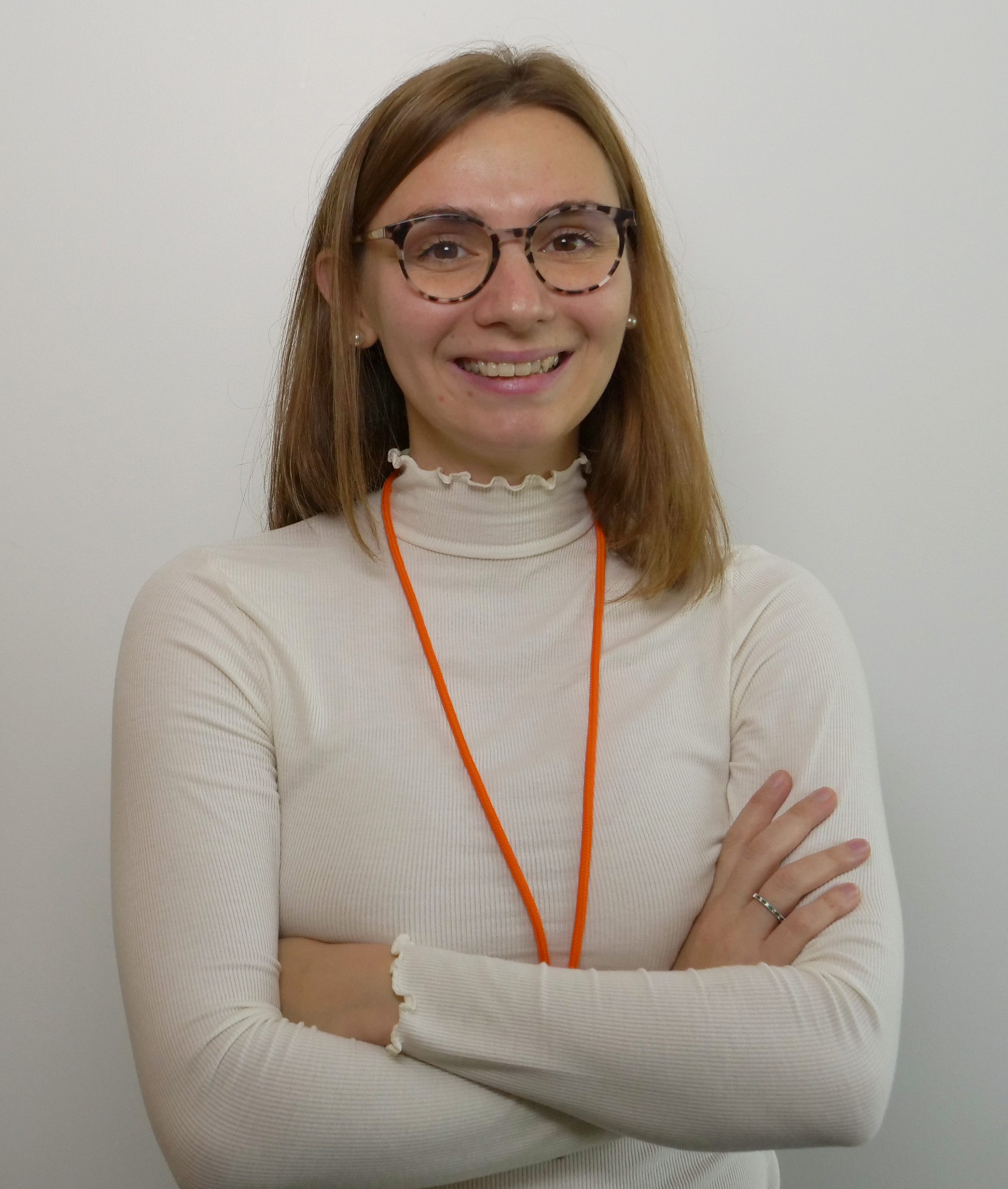 Femme souriante avec des lunettes, portant un haut à col montant et un collier orange, les bras croisés.