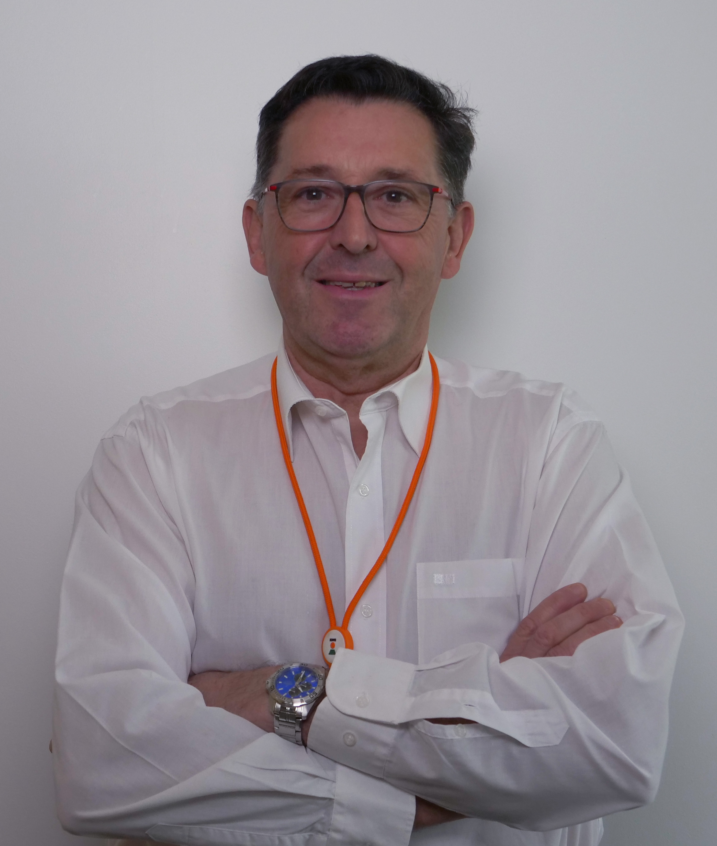 Homme souriant avec des lunettes, portant une chemise blanche et un collier orange, les bras croisés.