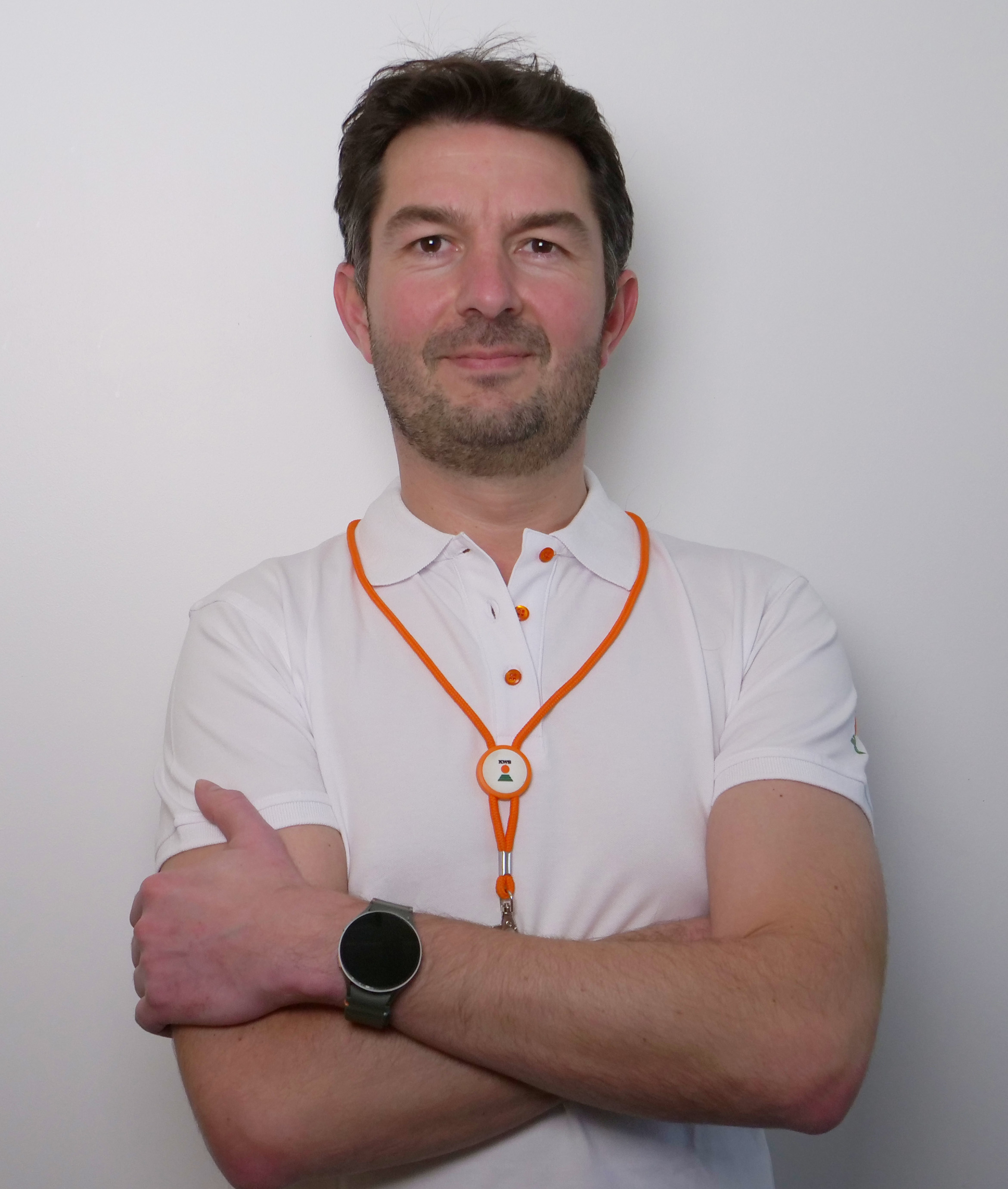 Homme avec un polo blanc et un collier orange, les bras croisés, se tenant devant un fond blanc.