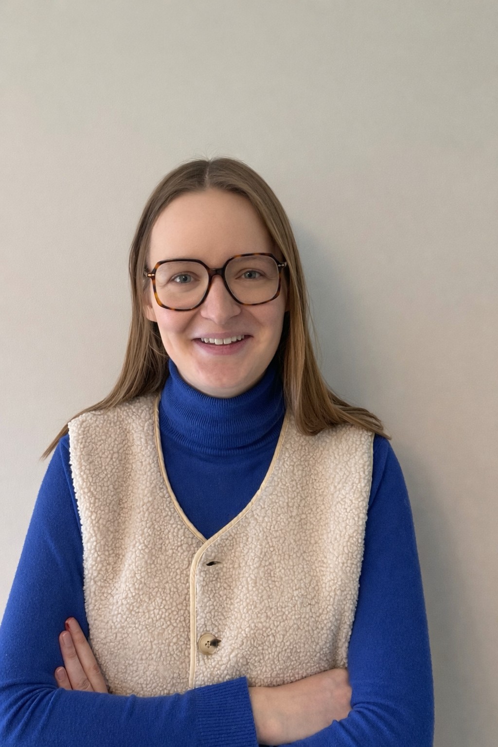 Portrait d'une femme souriante avec des lunettes, portant un pull bleu et un gilet beige. Elle se tient devant un mur clair.