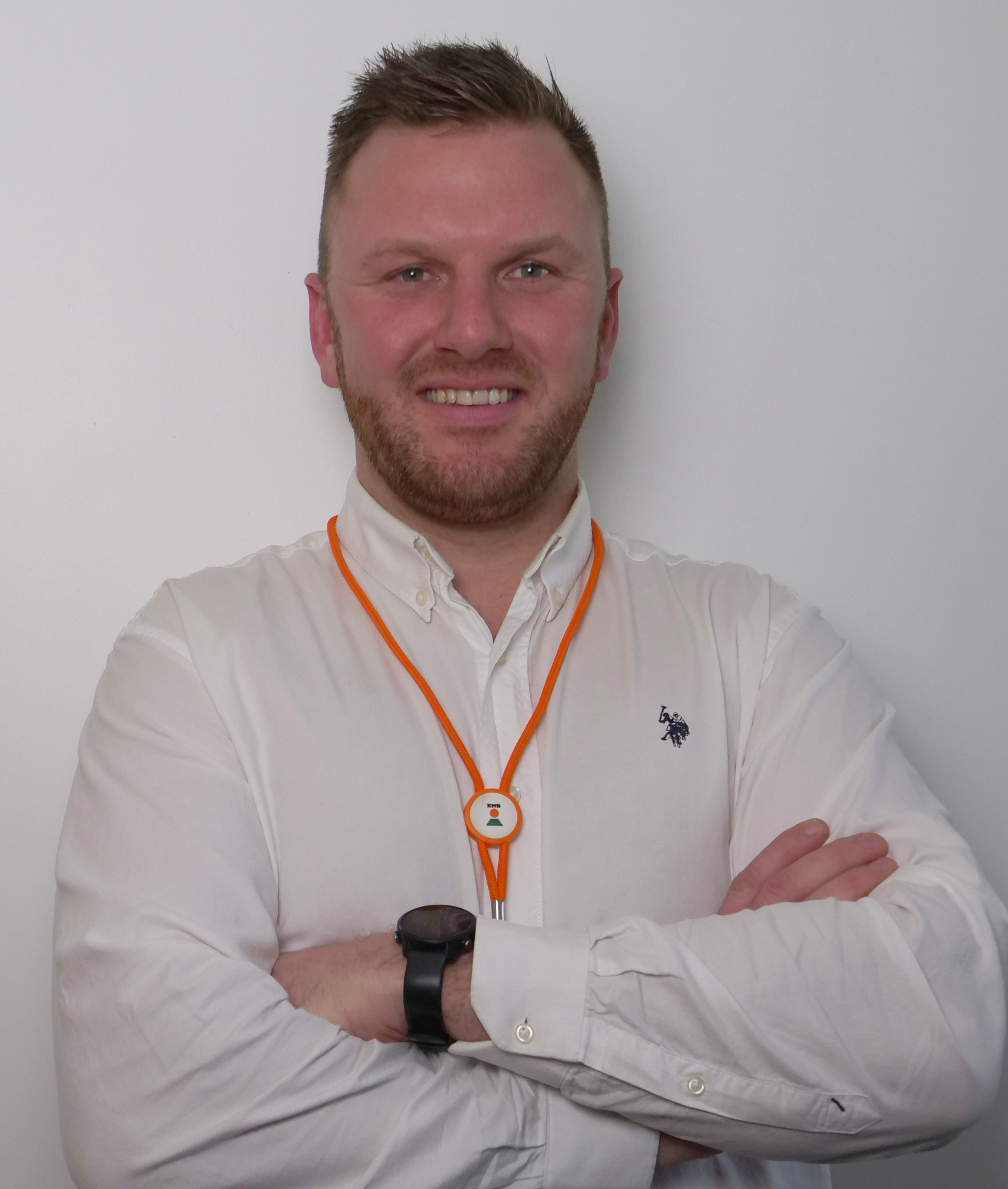Homme souriant avec une chemise blanche et un collier orange, les bras croisés. Fond neutre.